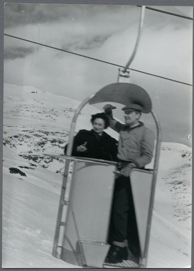 Ein Schwarz-Weiß-Foto eines Mannes und einer Frau auf einem Skilift, wobei der Mann eine Mütze trägt und die Frau Schuhe trägt, vor einem schneebedeckten Hintergrund mit dem Himmel oben.