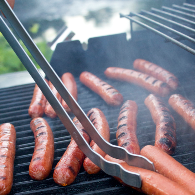 Würstchen auf einem Grill mit Zange, einer Person Hand und Pflanzen im Hintergrund bei Tageslicht.