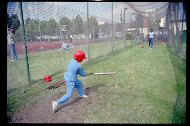 Ein junger Junge in einem blauen Kleid und einem roten Helm schwingt einen Baseballschläger auf einem grünen Feld mit verstreuten Schlägern, Bäumen, Gebäuden und einem klaren blauen Himmel im Hintergrund.
