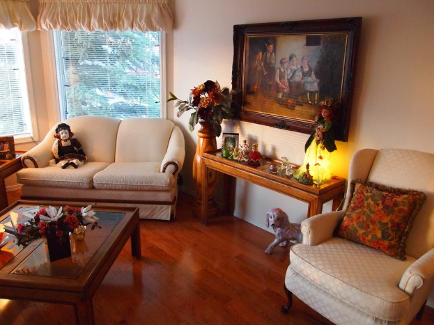 Eine Wohnzimmerszene mit zwei Sofas, eines mit einem Spielzeug und das andere mit einem Kissen, ein Tisch mit einem Spielzeug, einem Bild und verschiedenen Gegenst├Ąnden, eine Vase mit Blumen neben dem Tisch, ein Bild an der Wand, ein Fenster mit einem Vorhang und ein Spielzeug unter dem Tisch.