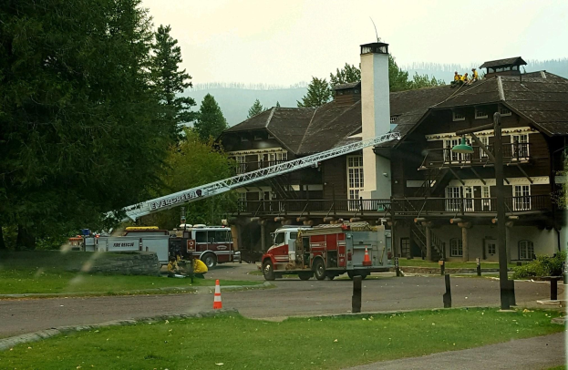Feuerwehrfahrzeug vor einem großen Gebäude mit Fenstern, Geländern, Säulen und einem Schornstein geparkt, umgeben von Absperrbaken, Gras, Bäumen und Laternen, mit einigen Menschen und Hügeln im Hintergrund unter einem klaren blauen Himmel.