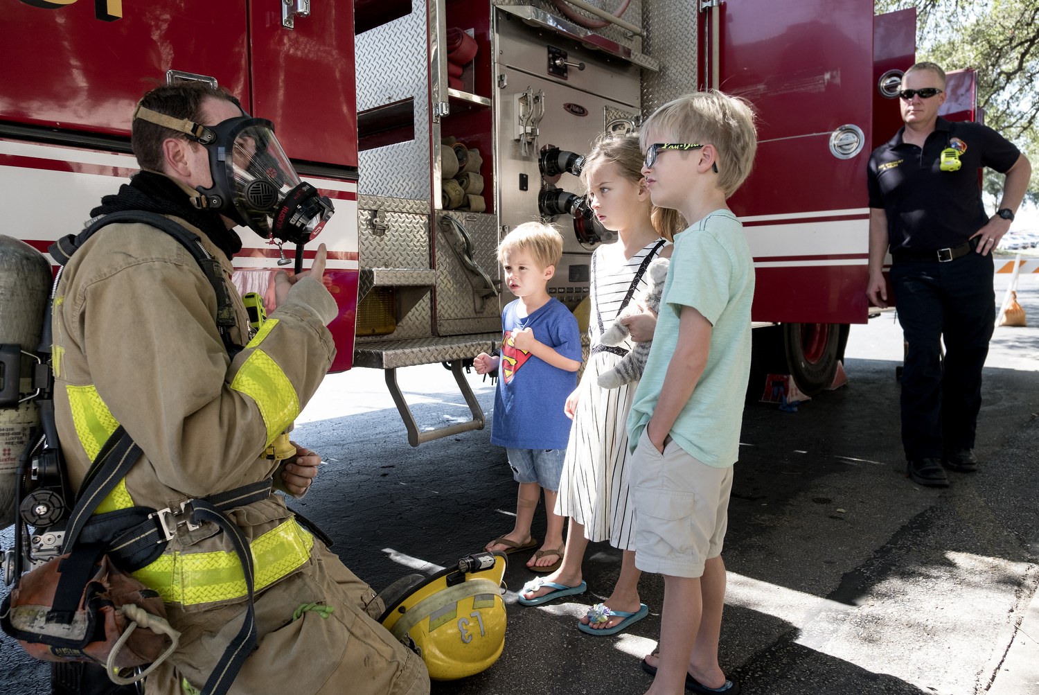 Ein Feuerwehrmann in einer Gasmaske spricht zu einer Gruppe von Kindern vor einem Feuerwehrauto, mit Bäumen und einem blauen Himmel im Hintergrund.