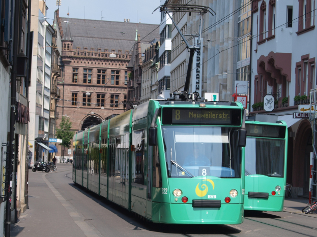 Zwei grüne Straßenbahnen fahren auf einer Straße mit hohen Gebäuden, Fahrräder auf dem Gehweg geparkt, Fußgänger auf der Straße, ein Baum im Hintergrund und ein klarer blauer Himmel.