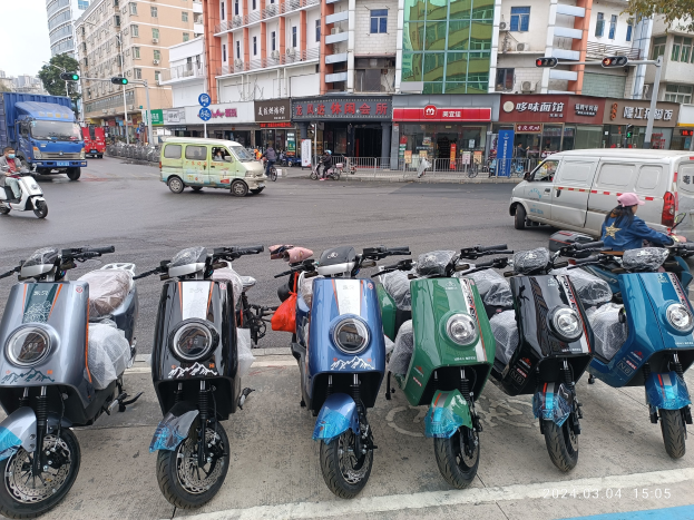 Eine Reihe von Elektroscootern, die auf einer Straße geparkt sind, mit einer Person in einer Mütze davor, vor einer Kulisse von Gebäuden, Ampeln, Bäumen und einem klaren blauen Himmel.