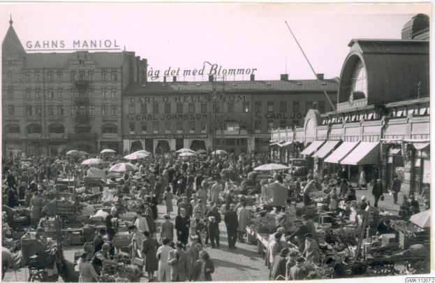 Schwarzes und weißes Foto eines belebten Berliner Markts mit Menschen, Gemüsewagen und Gebäuden mit Schildern im Hintergrund.