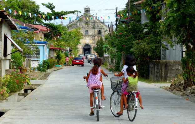 Drei Kinder fahren Fahrräder auf einer von Bäumen gesäumten Straße mit Gebäuden auf beiden Seiten, einem Auto und einem Gebäude im Hintergrund und Himmel darüber.