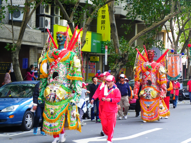 Eine Gruppe von Menschen in farbenfrohen Kostümen und Mützen marschiert auf einer Straße in einem chinesischen Neujahrsfest, mit Fahrzeugen, Bäumen, Gebäuden mit Fenstern und Klimaanlagen sowie Texttafeln im Hintergrund.