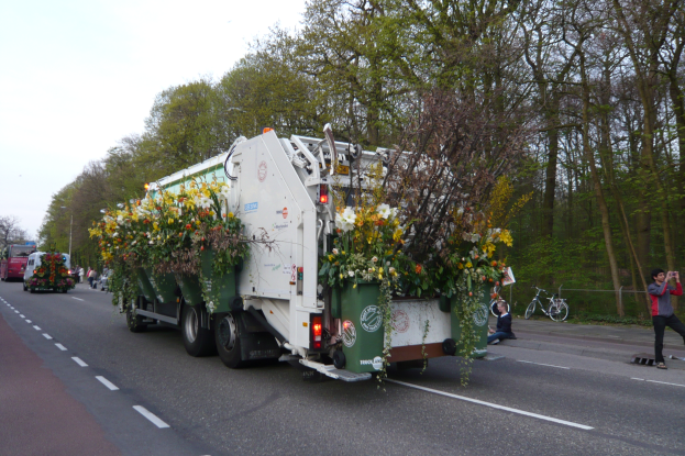 Ein Müllfahrzeug mit Blumen auf der Ladefläche fährt eine Straße entlang, umgeben von Bäumen und einem klaren blauen Himmel, mit ein paar Menschen und Fahrrädern im Hintergrund.