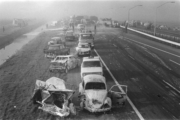 Ein Schwarz-Weiß-Foto von mehreren verunglückten Autos am Straßenrand bei Nacht, mit Straßenlaternen, einigen nahestehenden Menschen, Bäumen und einem Gewässer im Hintergrund.