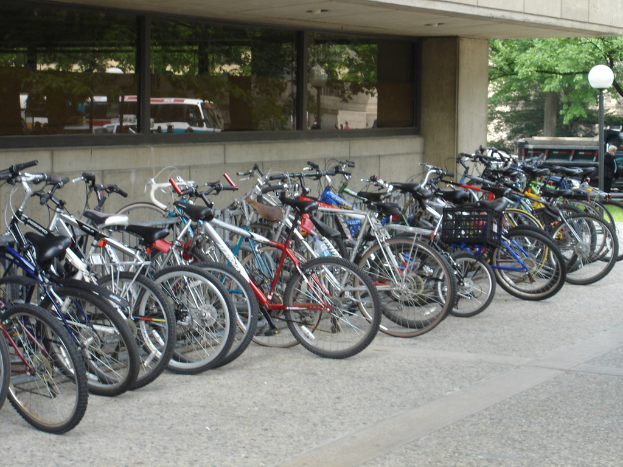 Mehrere Fahrr√°der sind auf einem Weg neben einer Laterne geparkt, mit einer Person auf der rechten Seite, Fahrzeugen, B√§umen und einem Geb√§ude im Hintergrund.