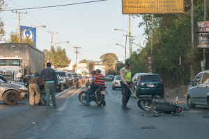 Eine Gruppe von Menschen steht um ein verunglücktes Motorrad auf der Straße herum, mit mehreren Fahrzeugen, darunter ein Lastwagen, und einem Hintergrund aus Bäumen, Pfählen, Lampen und Schildern unter dem Himmel.