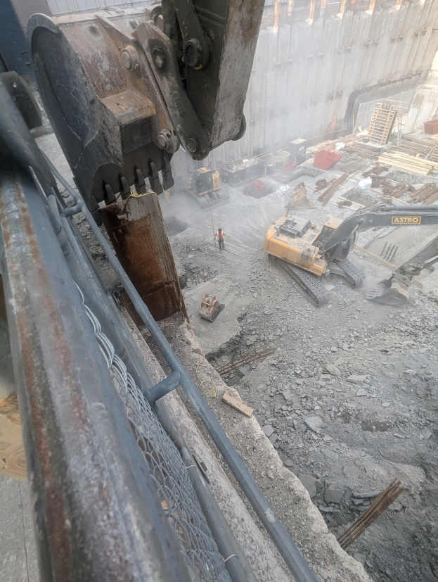 Großer Bagger auf einer Abrissstelle mit einem Gebäude im Hintergrund, mehreren Fahrzeugen, Personen und einem Metallobjekt links.