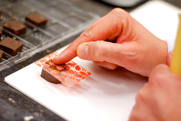 Eine Hand legt eine dekorative Abdeckung auf eine Schokolade, mit weiteren Schokoladen auf einem Tablett links und einem Tisch darunter.