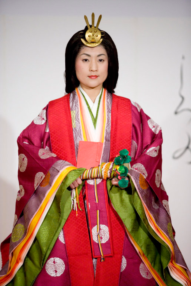 Eine Frau in einem traditionellen japanischen Kimono, das ein leuchtend rotes und grünes Kleid mit kunstvollen Mustern trägt, posiert selbstbewusst auf einem Laufsteg, ihr Haar zu einem ordentlichen Knoten gebunden.