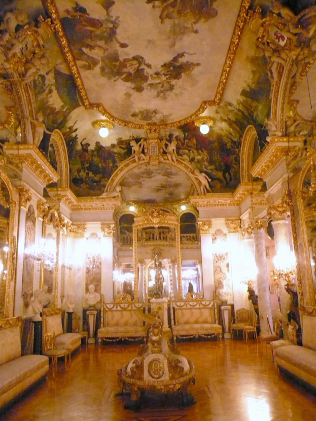 Ein großer, prunkvoller Raum voller verschiedener Möbel, Kronleuchter, Lampen und Gemälde, mit einer kunstvoll gestalteten Decke.