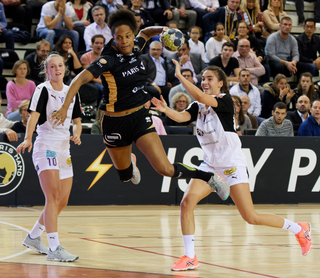 Eine Gruppe von Frauen beim Handballspiel auf einem Platz, mit einem Ball in der Mitte und Zuschauern und einem Banner im Hintergrund.