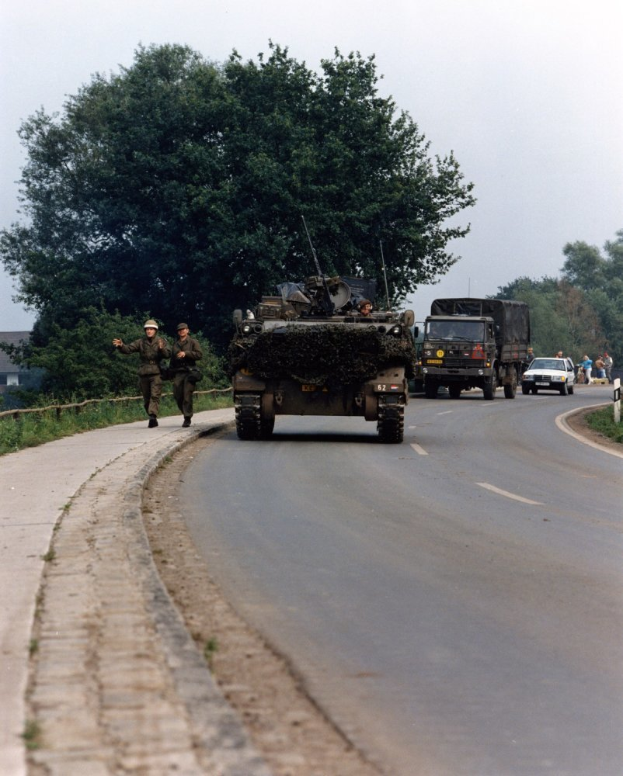 Ein militärisches Fahrzeug fährt auf einer Straße neben einem Wald, während uniformierte und helmtrugende Personen daneben hergehen, während im Hintergrund Gebäude und ein klarer blauer Himmel zu sehen sind.