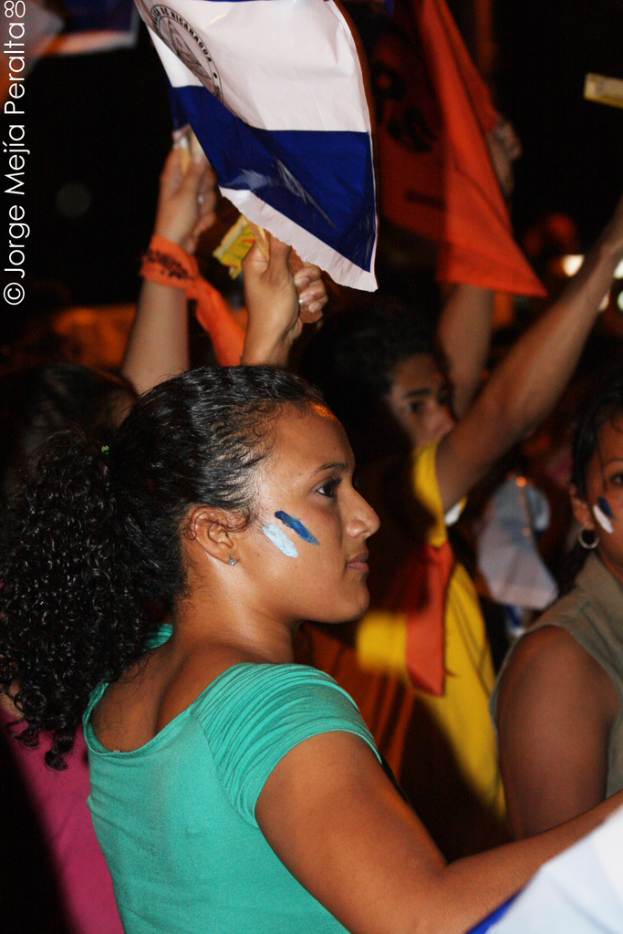 Eine Frau in einem grünen T-Shirt steht auf einer Demonstration und hält ein weißes Schild, mit vielen anderen Menschen hinter ihr.