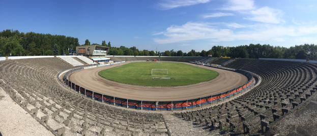 Ein leerer Stadion mit einem Fußballfeld, umgeben von Bäumen und einem Gebäude, unter einem bewölkten Himmel.