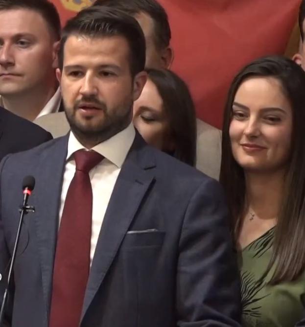 Ein Mann im Anzug spricht an einem Podium mit einem Mikrofon und blickt auf eine Menge hinter einem Banner, wahrscheinlich diskutiert er ein Regierungsverbot der sozialen Medien.