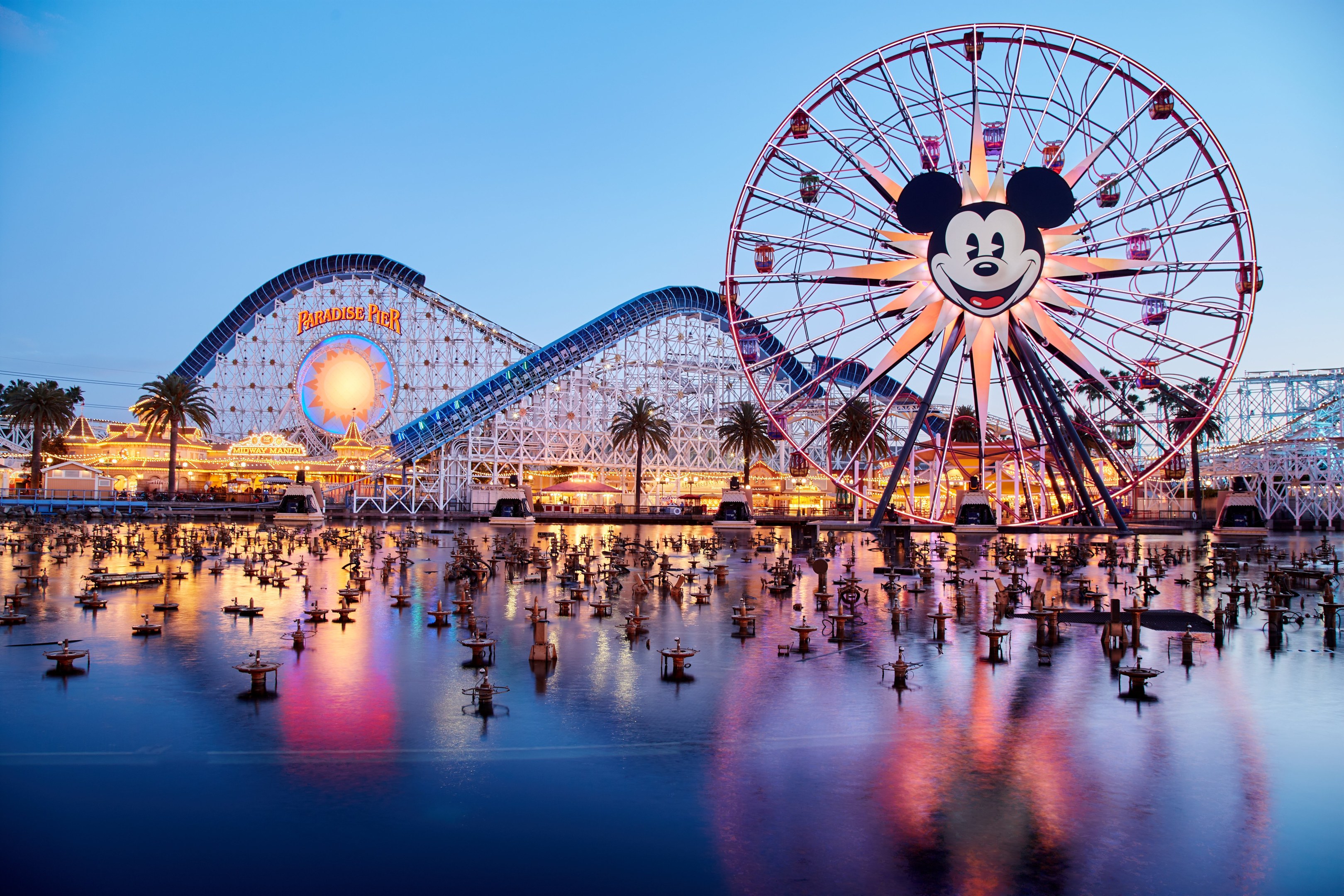 Eine lebendige Dämmerungsszene im Disneyland California Adventure mit einem riesigen Mickey-Maus-Rad im Vordergrund, einer Achterbahn im Hintergrund und Booten auf dem Wasser, mit einem farbenfrohen Himmel und silhouettenartigen Bäumen.