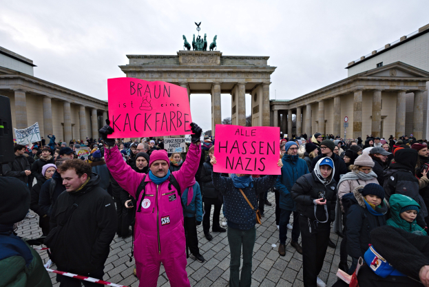 Eine Gruppe von Menschen mit Protestschildern vor dem Reichstagsgebäude in Berlin, mit Gebäuden und Statuen im Hintergrund unter einem klaren blauen Himmel.