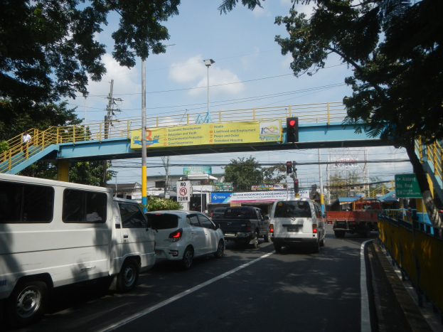Eine belebte Straße mit Fahrzeugen, eine Brücke mit Geländern und Treppen, Laternen, Verkehrszeichen, Schilder, Bäume, Gebäude und ein bewölkter Himmel.