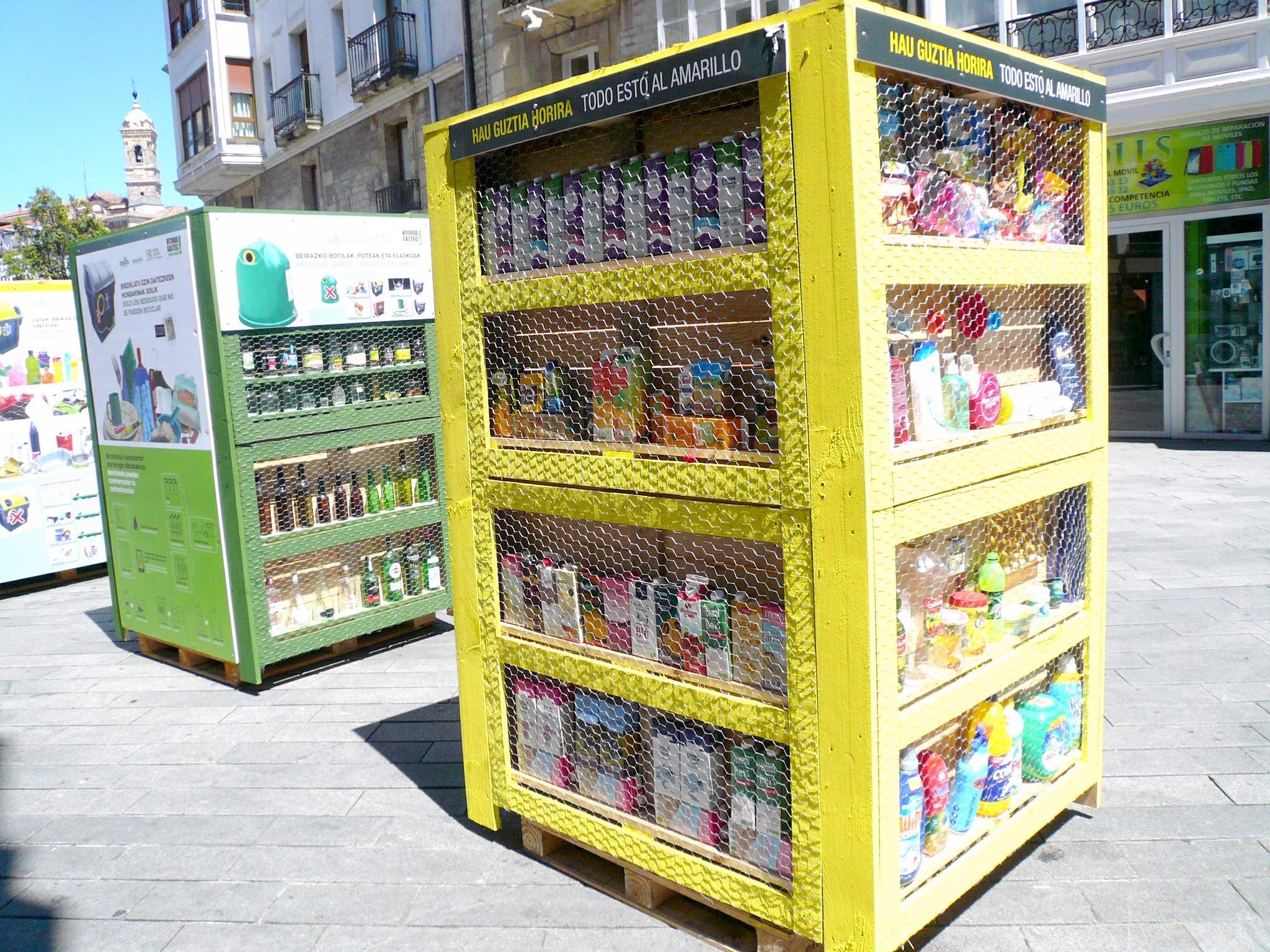 Ein gelber Verkaufsautomat auf einer Stadtstraße, gefüllt mit Gegenständen, die Ähneln Bauten und Bäumen, unter einem klaren blauen Himmel.