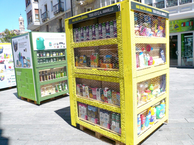 Ein gelber Verkaufsautomat auf einer Stadtstraße, gefüllt mit Gegenständen, die Ähneln Bauten und Bäumen, unter einem klaren blauen Himmel.