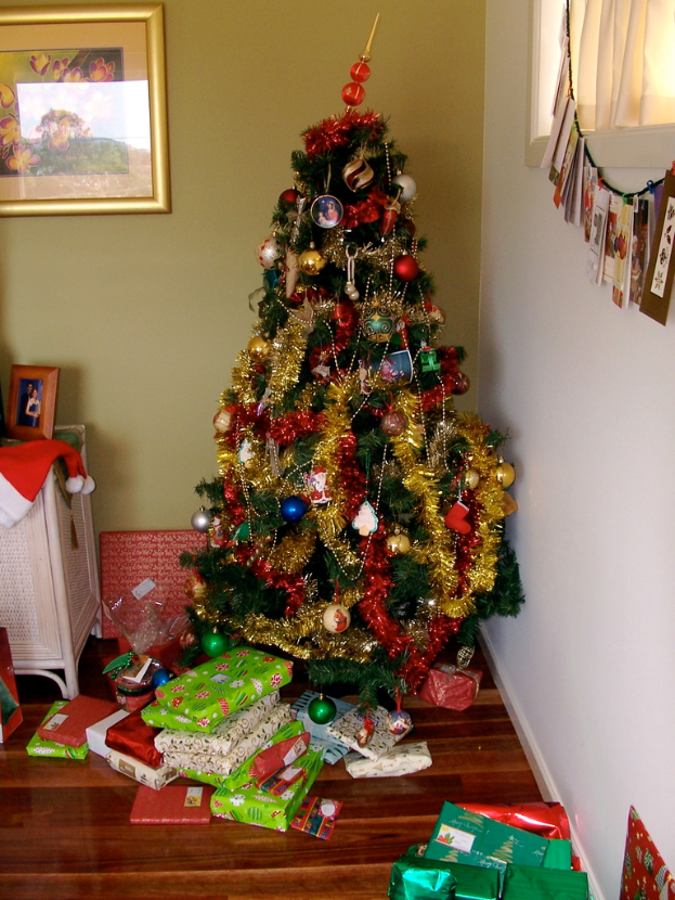 Ein geschmückter Weihnachtsbaum auf der rechten Seite mit Geschenken darunter, ein Foto auf der linken Wand und ein Vorhang am rechten Fenster.