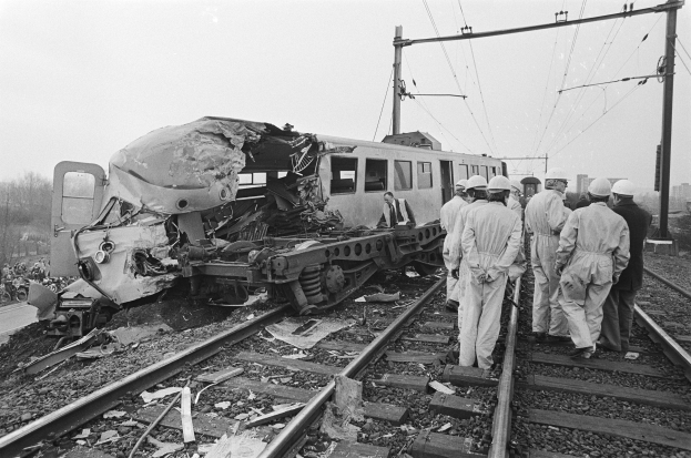 Ein Schwarz-Weiß-Bild einer Zugkollision auf den Schienen mit einer Gruppe von Helmträgern in der Nähe, vor einem Hintergrund aus Strommasten, Drähten, Bäumen, Gebäuden und einem klaren Himmel.