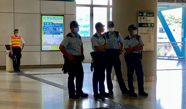 Eine Gruppe von Polizisten in Masken steht vor einem Gebäude mit Glastüren, Texttafeln, einer Säule und einem Geländer, mit Bäumen und Fahrzeugen im Hintergrund.