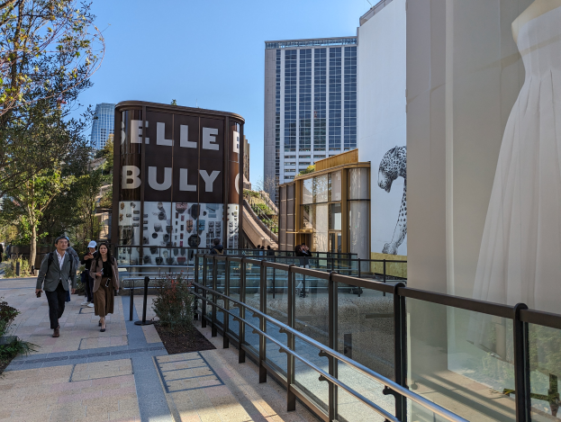 Menschen gehen eine Straße vor einem Gebäude mit einer Tafel, auf der "Elle Bully" steht, entlang, mit Bäumen und Pflanzen auf beiden Seiten, einer Schaufensterpuppe auf der rechten Seite und anderen Gebäuden und einem klaren blauen Himmel im Hintergrund.