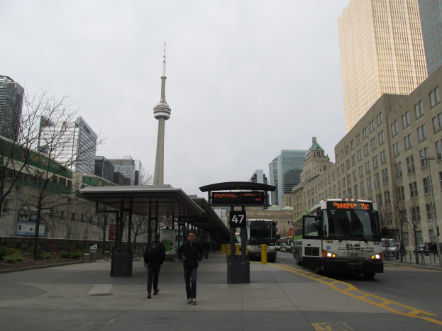 Eine belebte Stadtstraße mit einem Bus an einer Bushaltestelle, Fahrzeuge, Fußgänger, ein Schuppen, Bäume, Gebäude, ein Turm und der Himmel.