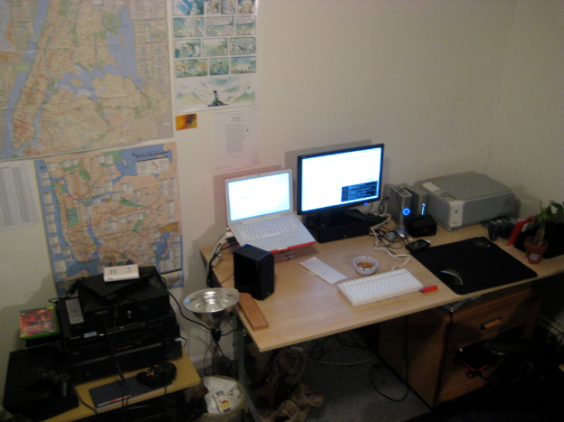 Ein Raum mit einem Tisch, auf dem ein Laptop, Lautsprecher, Monitor, Tastatur, Ball, Maus, Scanner und Papiere liegen, und einem weiteren Tisch mit Elektronikgeräten im Hintergrund, mit Diagrammen und Plakaten an der Wand dahinter.