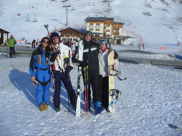 Vier Personen posieren für ein Foto mit Skating-Pads und Werkzeugen im Hintergrund, mit Gebäuden und anderen Menschen vor einer schneebedeckten Bergkulisse.