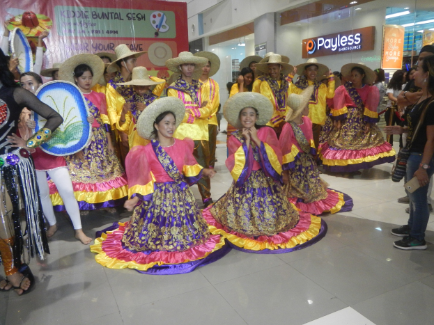 Eine Gruppe von Menschen in farbenfrohen Kleidern und Hüten, die in einem Einkaufszentrum während eines festlichen Ereignisses tanzen, während andere Musikinstrumente halten und ein Banner im Hintergrund zu sehen ist.