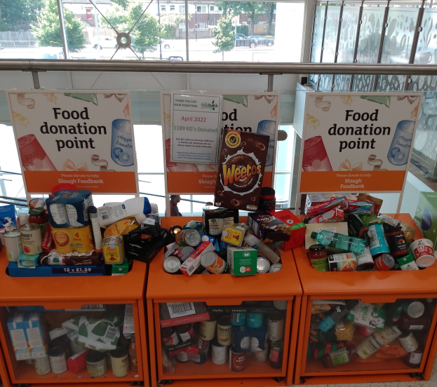 Orange Schübe mit Lebensmittelspenden, darunter Flaschen, Dosen und Kartons, mit informativen Schildern; außerhalb der Glasfenster sind Fahrzeuge, Bäume, Gebäude und Himmel zu sehen.