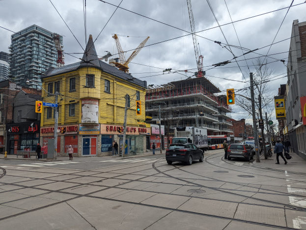 Eine Straßenszene in der Stadt mit einem gelben Gebäude im Bau, Autos, Fußgängern, Strommasten mit Drähten, Ampeln, Gebäuden mit Fenstern, Schildern, Bäumen, Kränen und einem bewölkten Himmel.