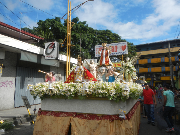 Ein Paradewagen mit Statuen, Blumen und Texttafeln, der eine Straße mit Laternenmasten, Gebäuden, Bäumen und einem bewölkten Himmel entlangfährt.