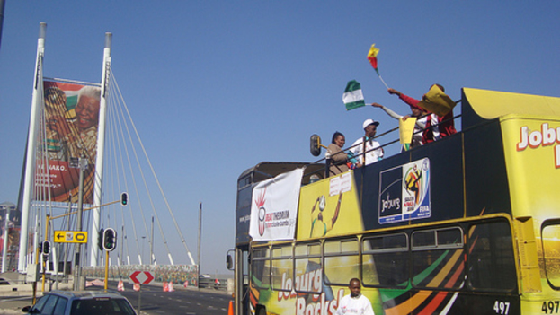 Ein Bus mit Menschen, die Fahnen schwenken, fährt auf einer Straße mit einem Auto links, einem Plakat in der Hintergrund und Verkehrsampeln und Masten entlang der Straße, unter einem klaren Himmel.