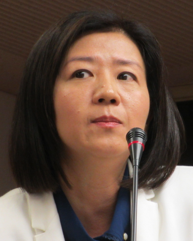 Eine Frau in einer weißen Jacke und einem Anzug spricht in ein Mikrofon vor einer Wand und hat einen ernsten Gesichtsausdruck.