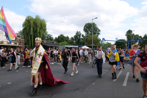 Eine Gruppe von Menschen marschiert bei der Christopher Street Day Parade 2018, einige mit Musikinstrumenten und andere mit Mützen, die eine Regenbogenflagge tragen, durch eine Straße mit Laternenpfählen, Bäumen, Hütten und einem bewölkten Himmel.