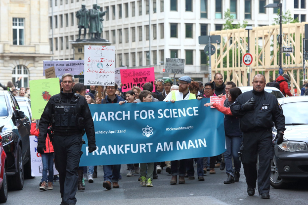 Gruppe von Menschen marschiert mit einem "March for Science Frankfurt am Main"-Schild die Straße entlang, mit Autos, die daneben fahren und Gebäude, Statuen, Laternen, Schilder und Bäume im Hintergrund.