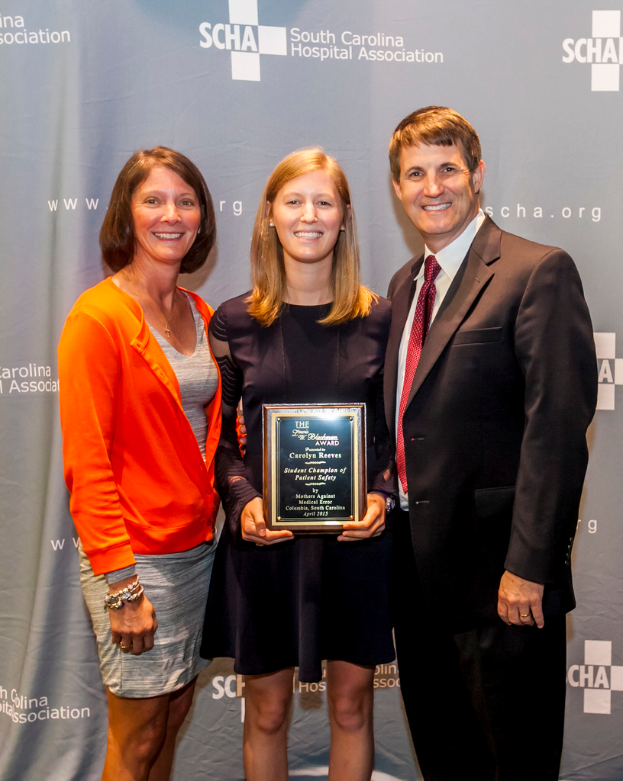 Drei Personen, zwei Frauen und ein Mann, die zusammenstehen, wobei eine Frau einen Preis hält, vor einem grauen Hintergrund.