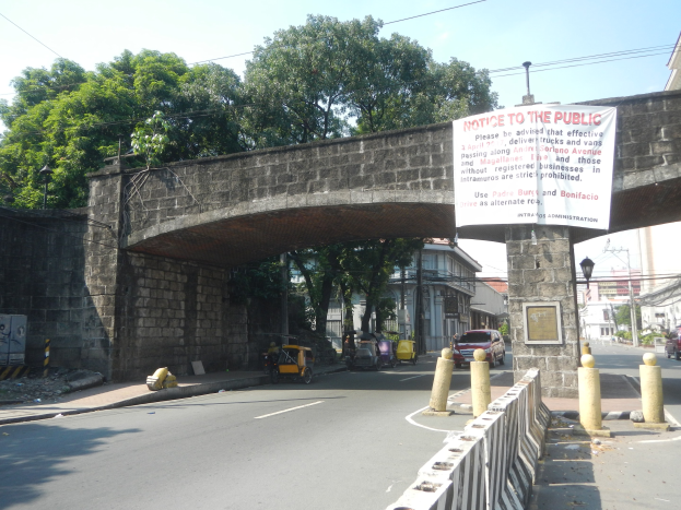 Öffentliche Bekanntmachung auf einer Brücke über einer Straße mit Fahrzeugen, einem Geländer auf der rechten Seite, Gebäuden, Bäumen, Strommasten, Drähten und Himmel im Hintergrund.
