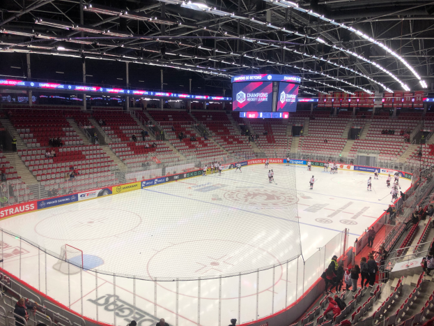Ein Hockey-Spiel in einer großen Arena mit Zuschauern auf den Sitzplätzen und Stehplätzen, einem Zaun um die Eisfläche, einem Display mit der Aufschrift 'St. Louis Blues vs Detroit Red Wings', Metallrahmen und einer Decke mit Deckenleuchten.