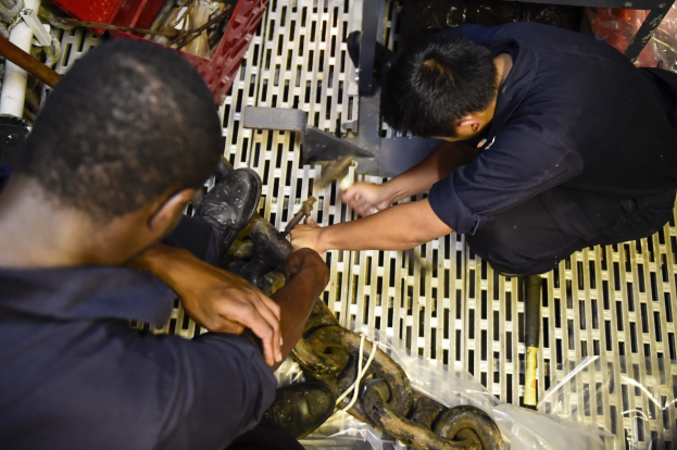 Zwei Männer in einer Fabrik, die an einer Maschine arbeiten und Werkzeuge halten, umgeben von Rohren und anderen Gegenständen.