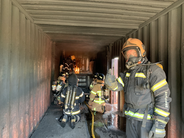 Eine Gruppe von Feuerwehrleuten in einem Seecontainer, mit Helmen, Handschuhen und Sauerstoffflaschen, einer Person mit einem Rohr und einem Feuerlöscher im Hintergrund.