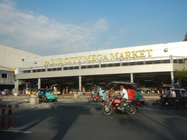 Eine belebte Straßenszene auf dem Pasig City Mega Market mit Fahrzeugen und Fußgängern, einem Gebäude mit Text im Hintergrund und einem bewölkten Himmel.